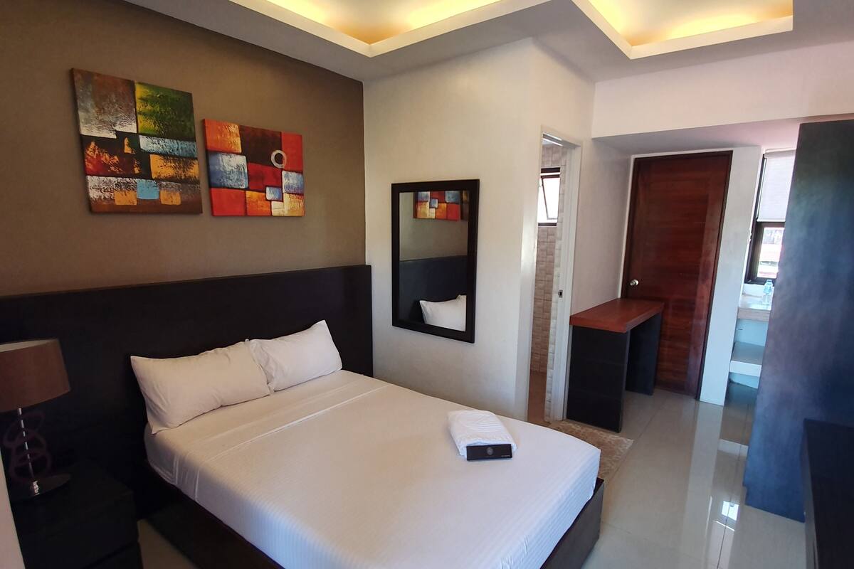 Una habitación de hotel moderna con una cama grande, una mesita de noche, una lámpara y dos cuadros abstractos en la pared.