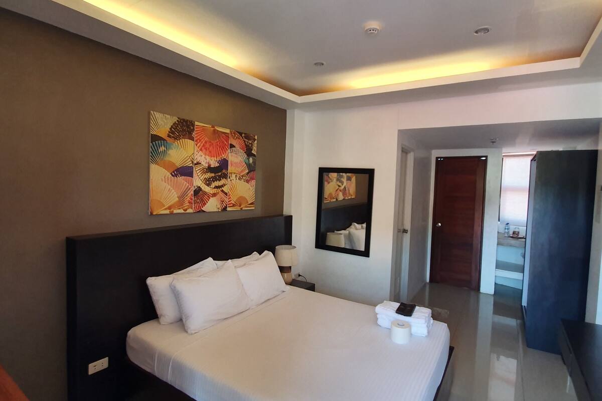 Una habitación de hotel moderna con una cama grande, cabecera, mesita de noche, lámpara y una obra de arte colgada en la pared.