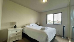 A hotel room with a bed, a nightstand with a lamp, a window with a view, and a radiator. - Seabreeze 6 - Opposite Tuncurry Bowling Club! (Tuncurry)