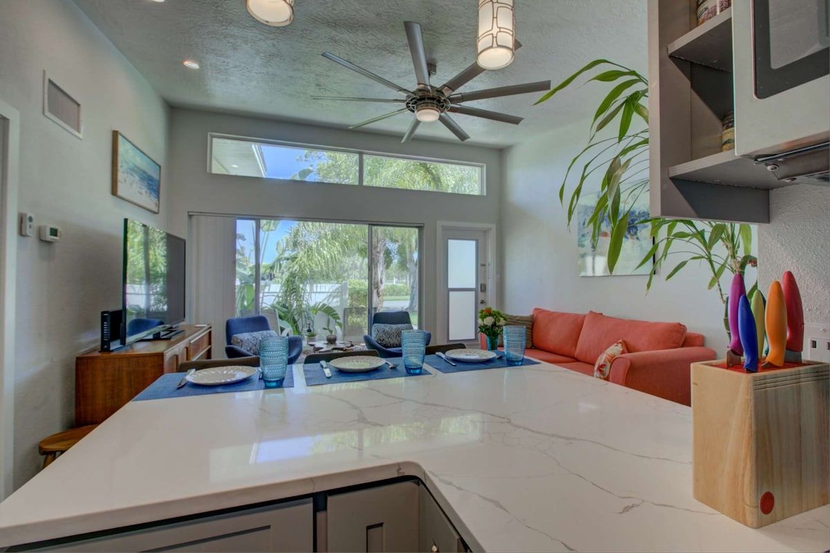 A modern kitchen with a large island, a ceiling fan, and a dining area with a red sofa and blue chairs.