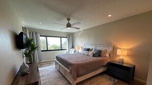 Une chambre à coucher comprenant un lit, des tables de chevet, un bureau, un ventilateur de plafond, une fenêtre avec des rideaux et une vue sur l’extérieur.