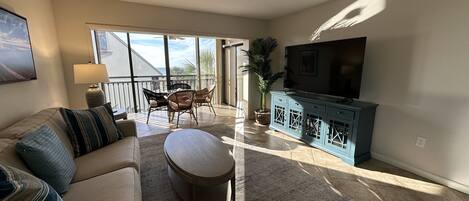Een ruime woonkamer met een bank, een voetenbankje en een televisie op een kast. Er is een balkon met uitzicht op de omgeving.