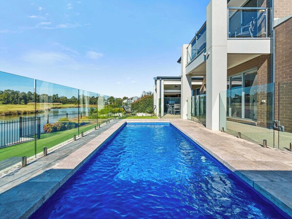 A modern pool with a clear view of the surrounding landscape, a glass fence, and a building with large windows and a balcony.