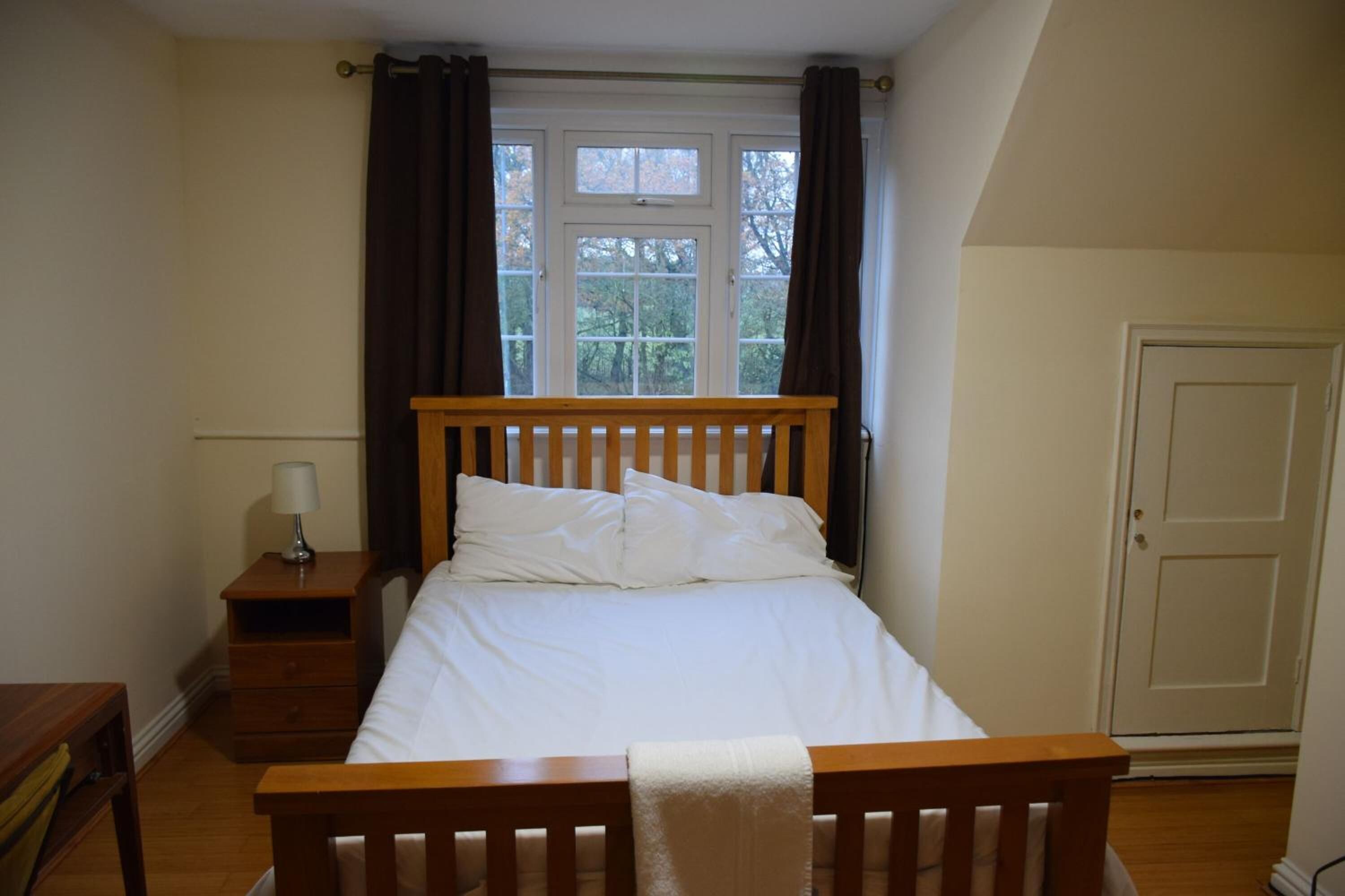 A bedroom with a wooden bed, a nightstand with a lamp, and a window with curtains.