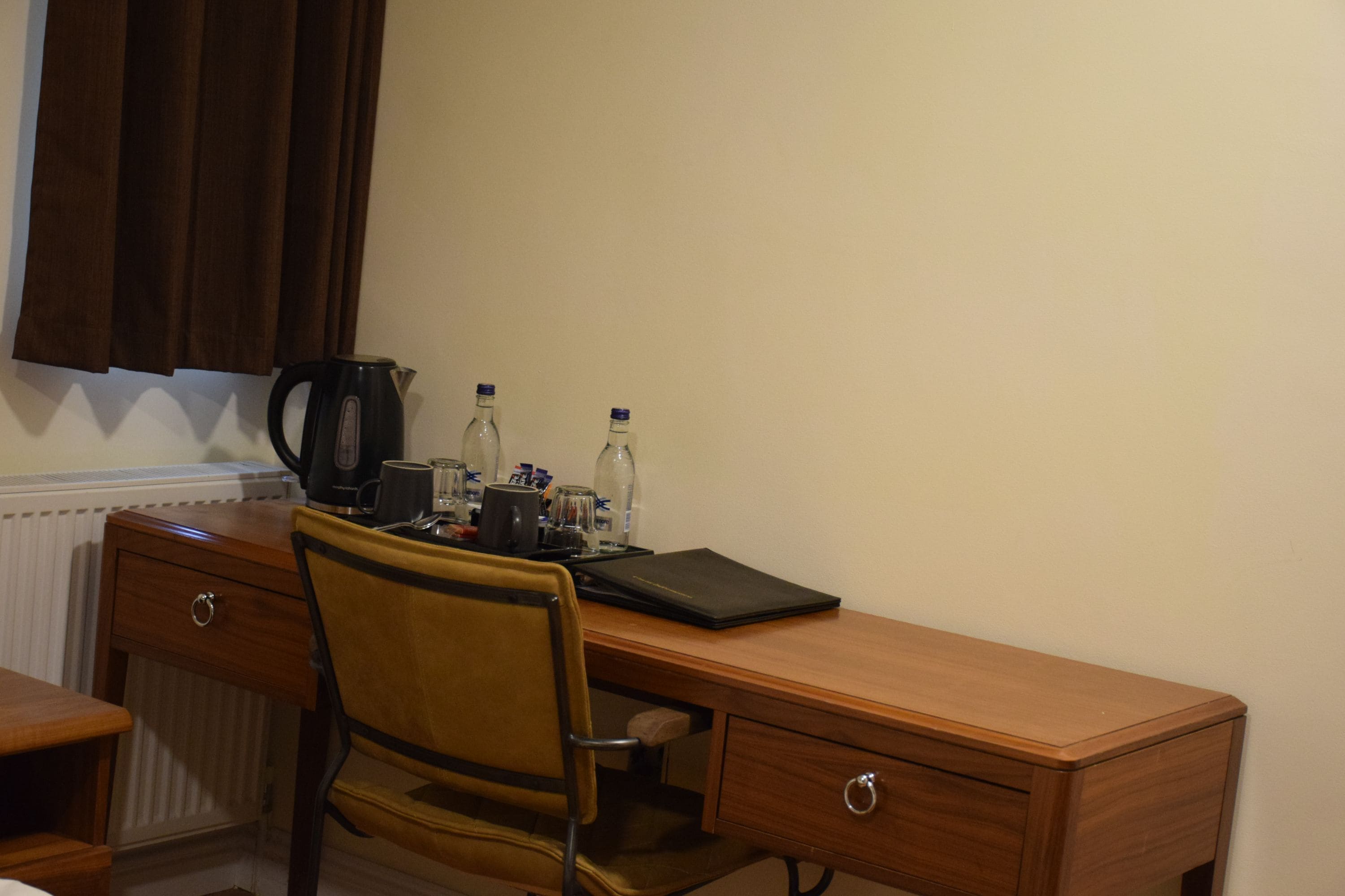 Un escritorio de madera con una silla, un radiador, un ventanal con cortinas marrones y varios objetos sobre el escritorio, incluyendo una carpeta negra, un vaso y botellas.