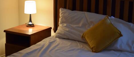 A bed with white bedding and a wooden headboard, a nightstand with a lamp, and a single yellow pillow.