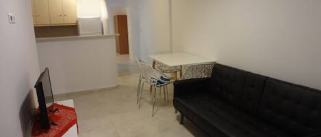A compact living space with a black sofa, a small dining table with chairs, a TV on a stand, and a kitchenette with cabinets.