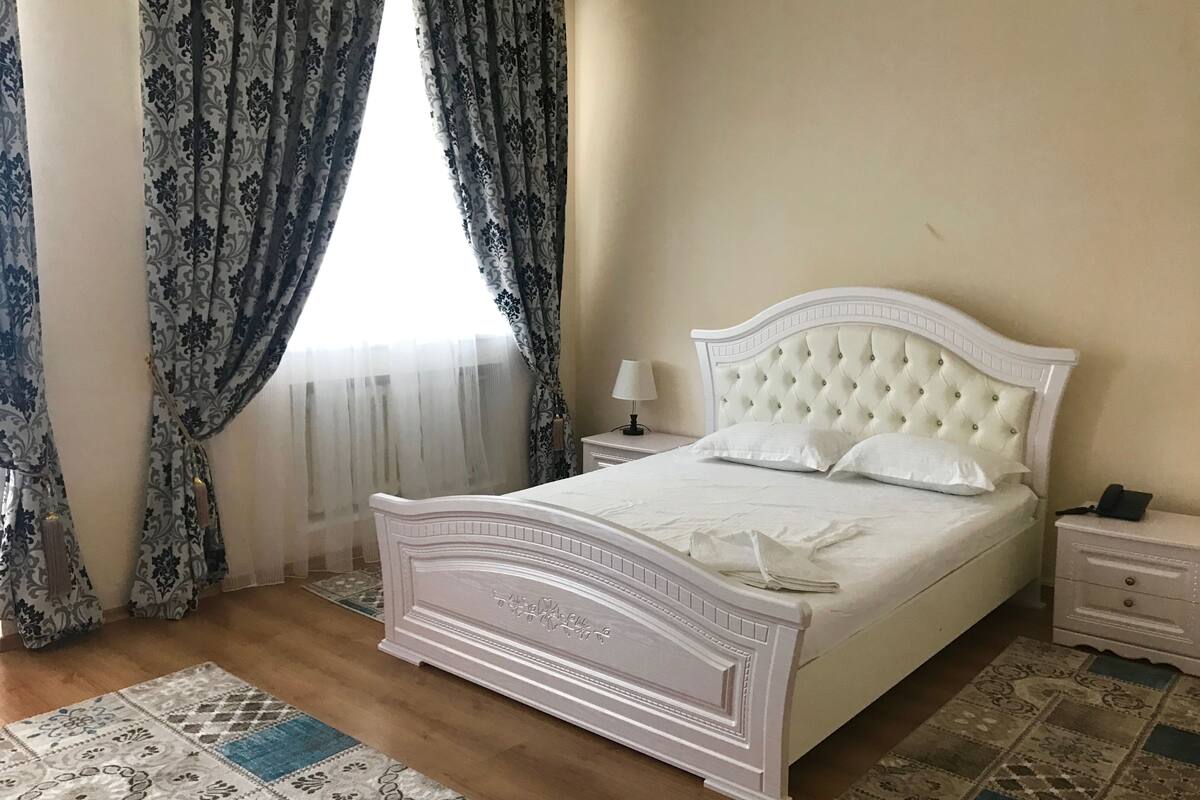 Un dormitorio con una cama blanca, una mesita de noche con lámpara y alfombras estampadas en el suelo.