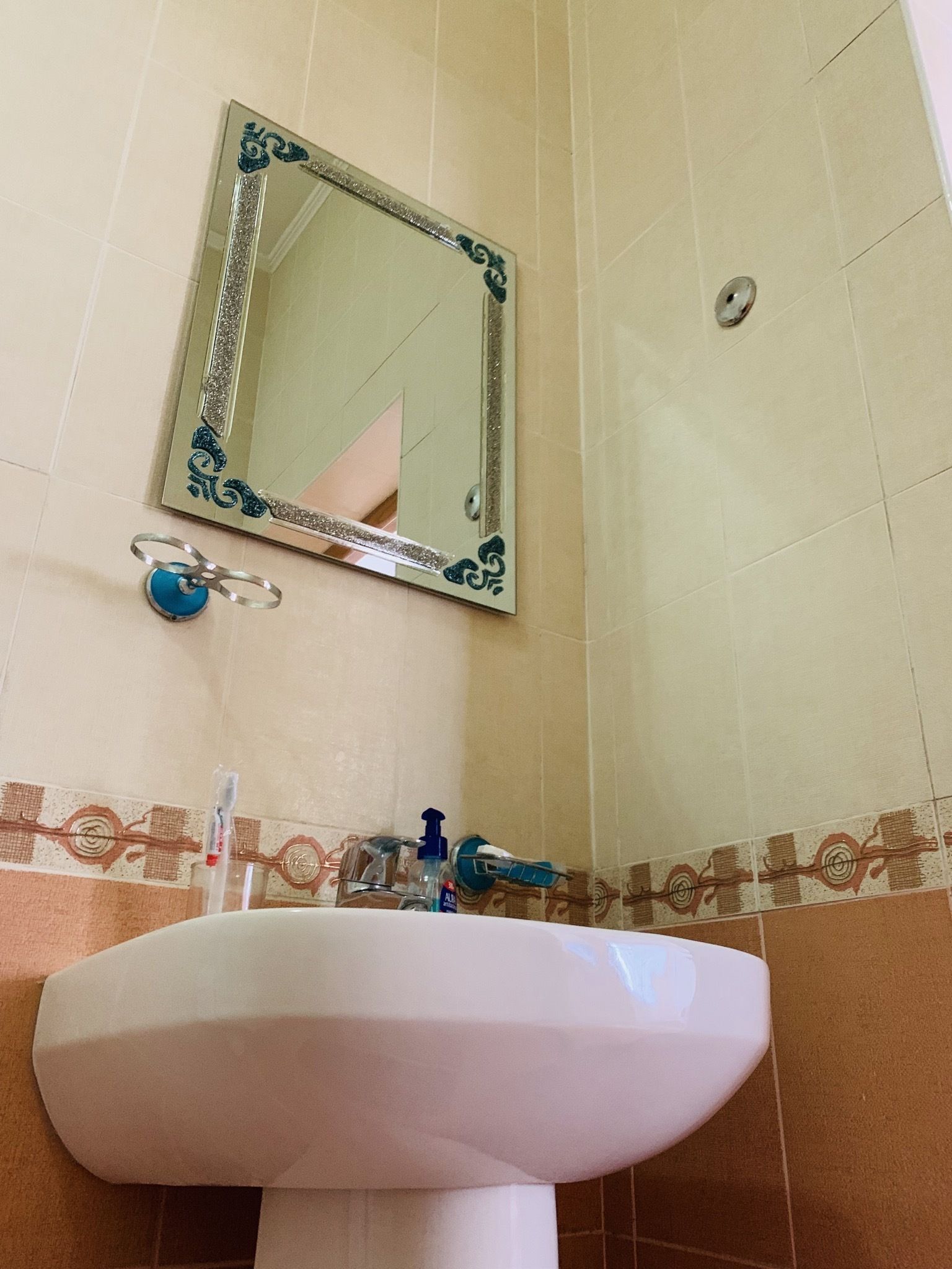 Une salle de bain avec un lavabo blanc, un miroir doté d’un cadre décoratif et un mur carrelé avec une bordure à motifs.
