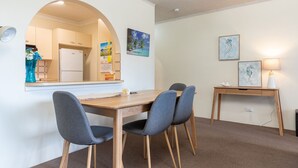 A dining area with a wooden table, two grey upholstered chairs, a small wooden side table with a lamp, and a mirror reflecting a kitchen area. - Seahorse 4 - Ground floor, and in the most requested area of Tuncurry! (Tuncurry)