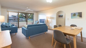 A modern living room with a sofa, a dining table, and chairs. There is a TV and a lamp. - Seahorse 4 - Ground floor, and in the most requested area of Tuncurry! (Tuncurry)