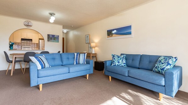 A living room with two blue sofas, a dining table with chairs, a kitchen area, and a painting on the wall. - Seahorse 4 - Ground floor, and in the most requested area of Tuncurry! (Tuncurry)