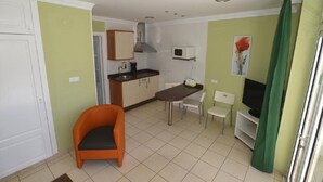 A compact kitchen with a microwave, stove, and sink. There is a dining area with a table and chairs, and a small orange chair. A television is on a stand to the right. - Apartamentos Arco Iris (San Bartolomé de Tirajana)