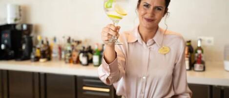 A person holding a glass with a lemon slice, standing in a kitchen with a countertop filled with various bottles.