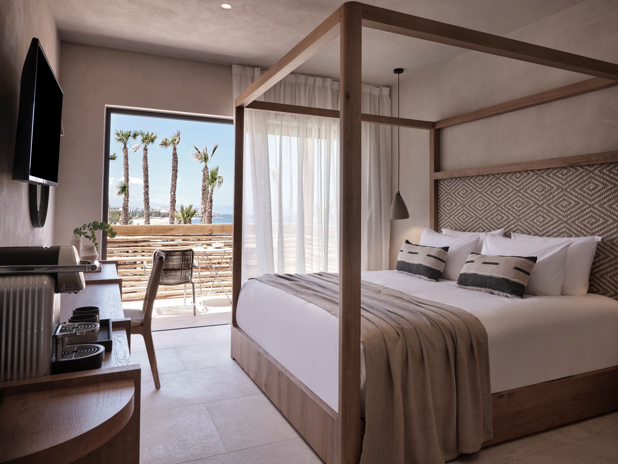 A bedroom with a four-poster bed, a desk with a coffee machine, and a view of palm trees and a beach.