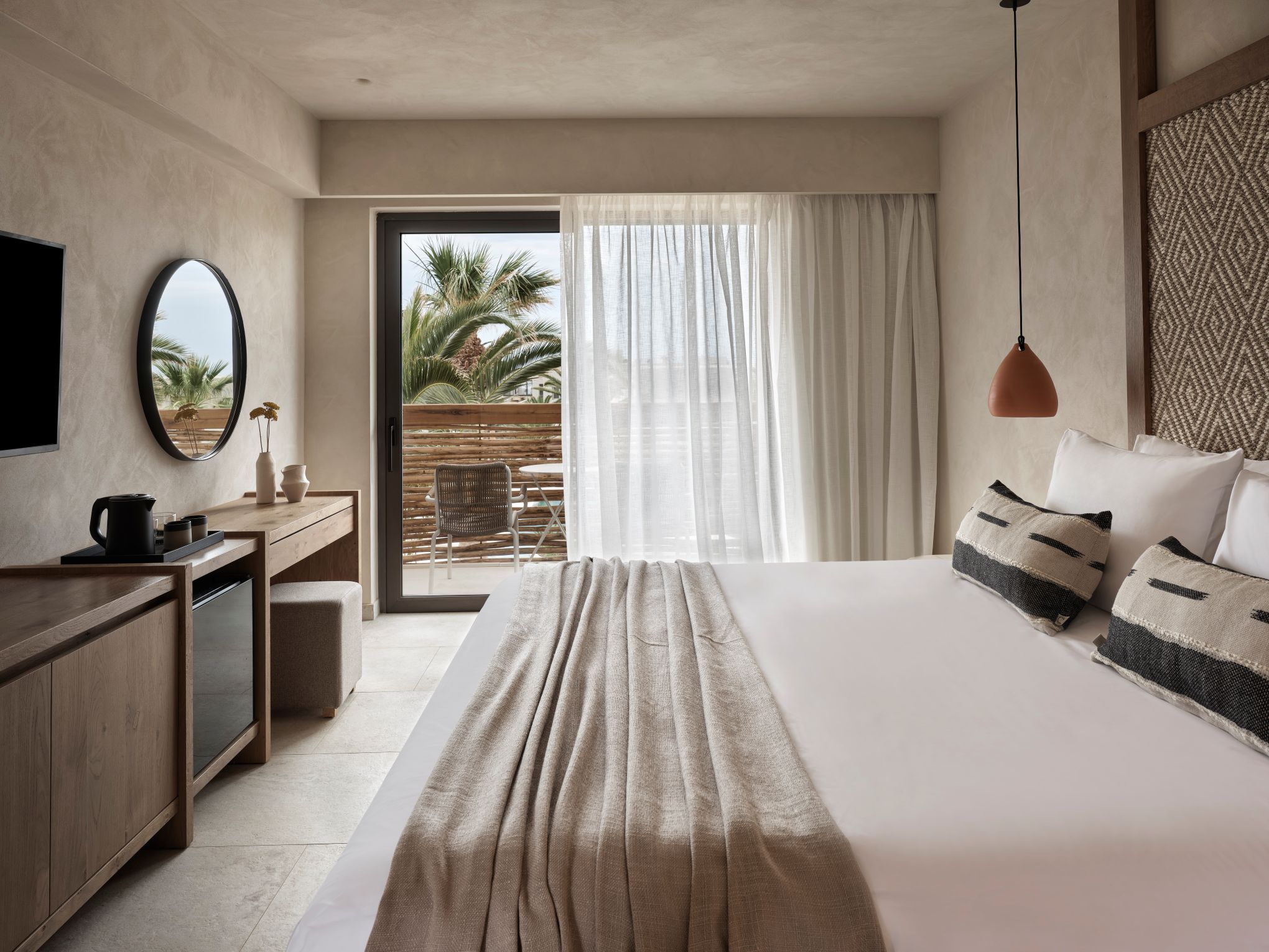 A modern hotel room with a large bed, a desk, a chair, a television, and a view of palm trees through the window.