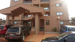 A multi-story building with a central entrance, balconies, and a parking area with cars. - MJ Appartments (Accra)