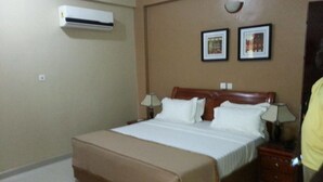 A hotel room with a bed, bedside lamps, a wall-mounted air conditioner, and two framed pictures on the wall. - MJ Appartments (Accra)