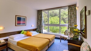 A hotel room with two beds, a desk, a chair, a window with curtains, a painting on the wall, and a vase with flowers. - Adriatica Rooms (Dubrovnik)