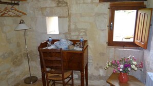 Une pièce rustique avec un mur en pierre, une table en bois ornée d’un vase de fleurs, un lampadaire et une fenêtre avec des volets en bois.