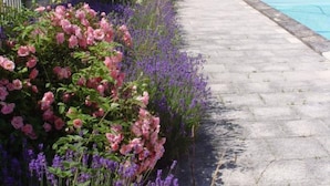 Un pont de pierre menant à une piscine, entouré de parterres fleuris où s’épanouissent des fleurs roses et violettes.