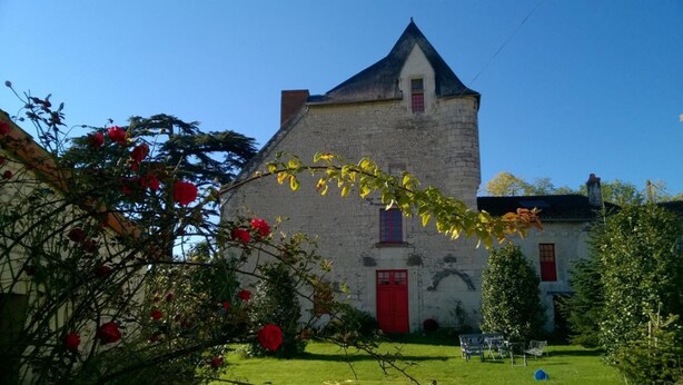 Un bâtiment en pierre avec une porte rouge et une petite fenêtre, entouré d’un jardin bien entretenu où poussent des rosiers.