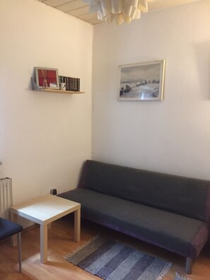 A living room with a grey sofa, a small white table, a bookshelf with books and a framed picture, and a radiator. - Zimmervermietung Lösken 2 (Duisburg)
