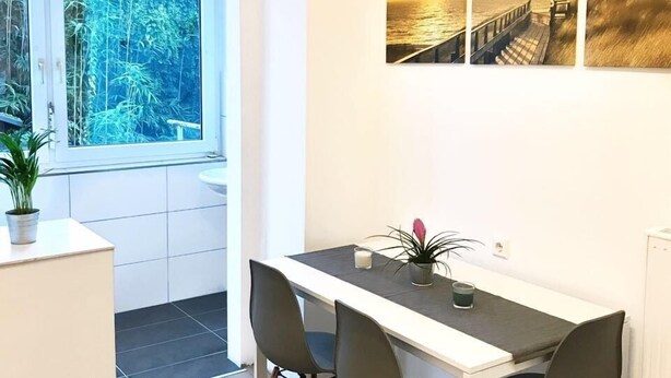 Uma cozinha moderna com bancada branca, uma mesa de jantar com quatro cadeiras e uma janela grande com vista para o verde.
