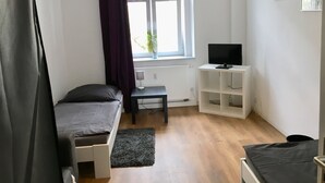 Quarto pequeno e bem iluminado com uma cama, uma escrivaninha com computador, uma lumin ária e uma televisão.
