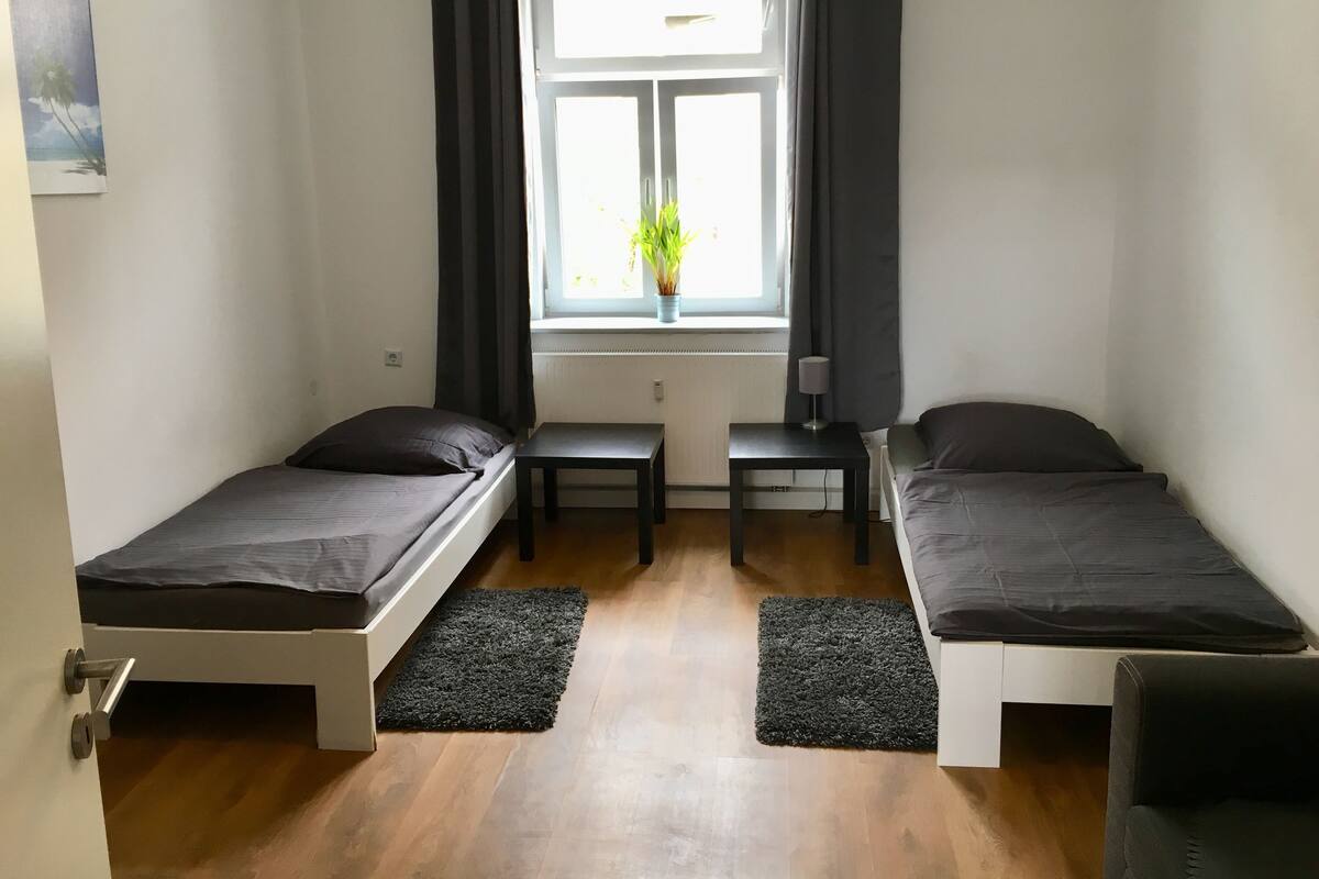 Ein kleiner, moderner Raum mit zwei Einzelbetten, einem kleinen Tisch, einem Fenster mit Vorhängen und einer Pflanze auf der Fensterbank.