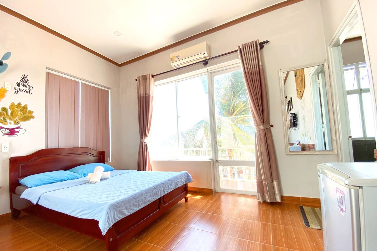 Un dormitorio con una cama de madera, un ventanal con cortinas, un refrigerador pequeño y un aire acondicionado instalado en la pared.