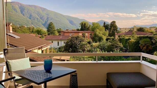 A balcony with a table, chairs, and a view of a mountainous landscape. - Bijou Lido Golf (Ascona)