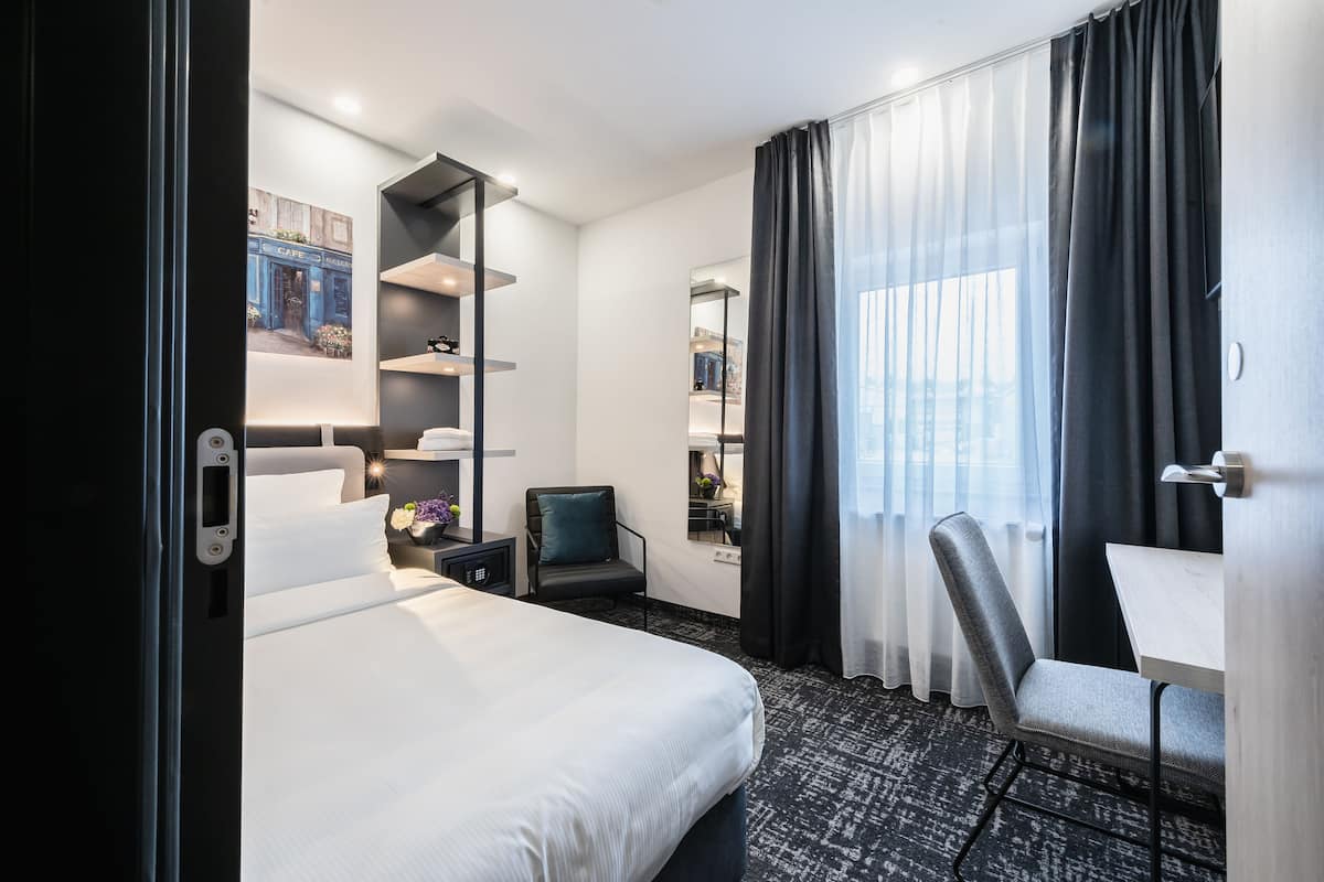 Hotellihuone, jossa on sänky, työpöytä, tuoli, kirjahylly ja suuri ikkuna, jossa on verhot.