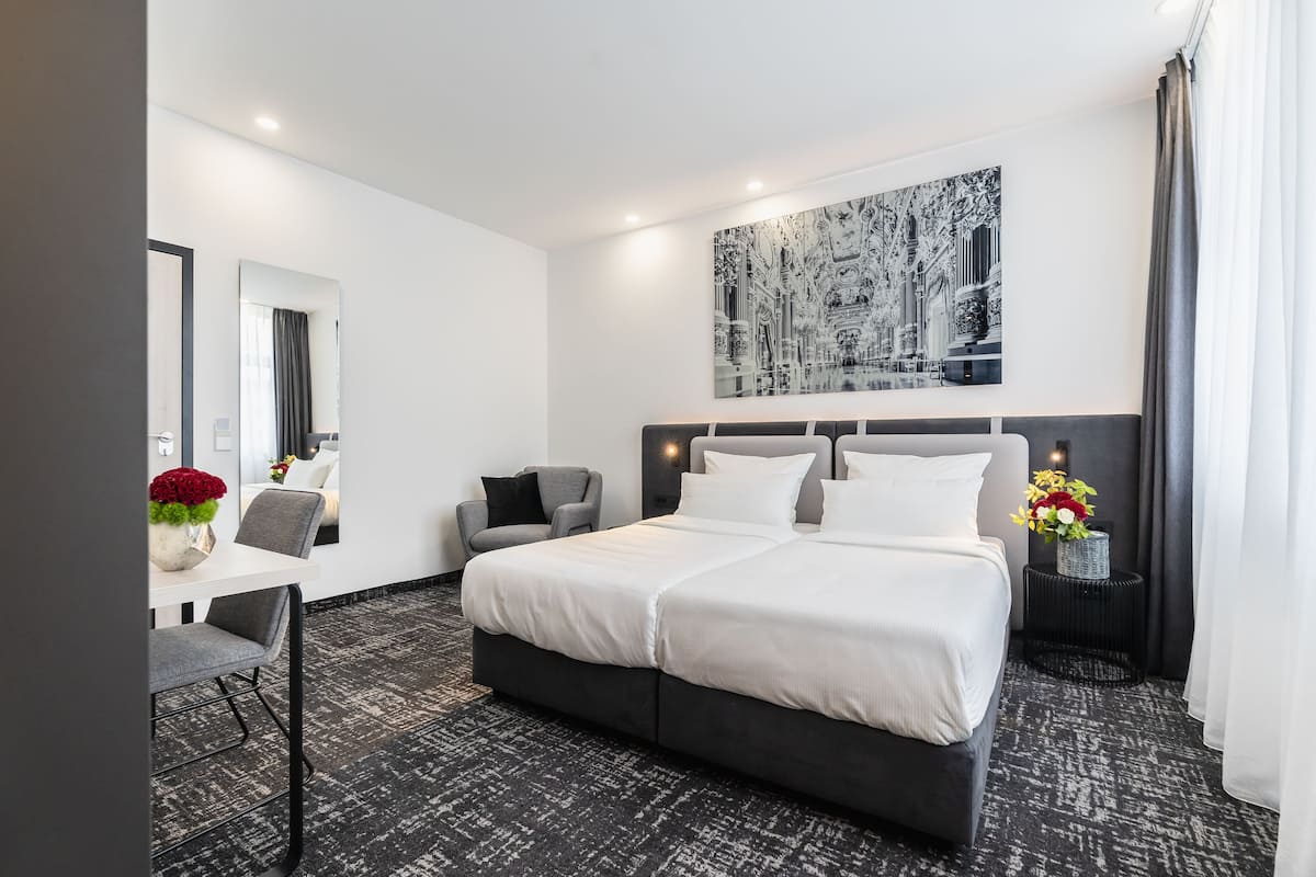 Moderni hotellihuone, jossa on suuri sänky, työpöytä tuolilla, pieni sivupöytä maljakko täynnä kukkia ja suuri kehystetty taideteos seinällä.