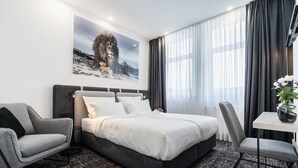 A modern hotel room with a large bed, a grey armchair, a small table with a vase, and a painting of a lion on the wall.