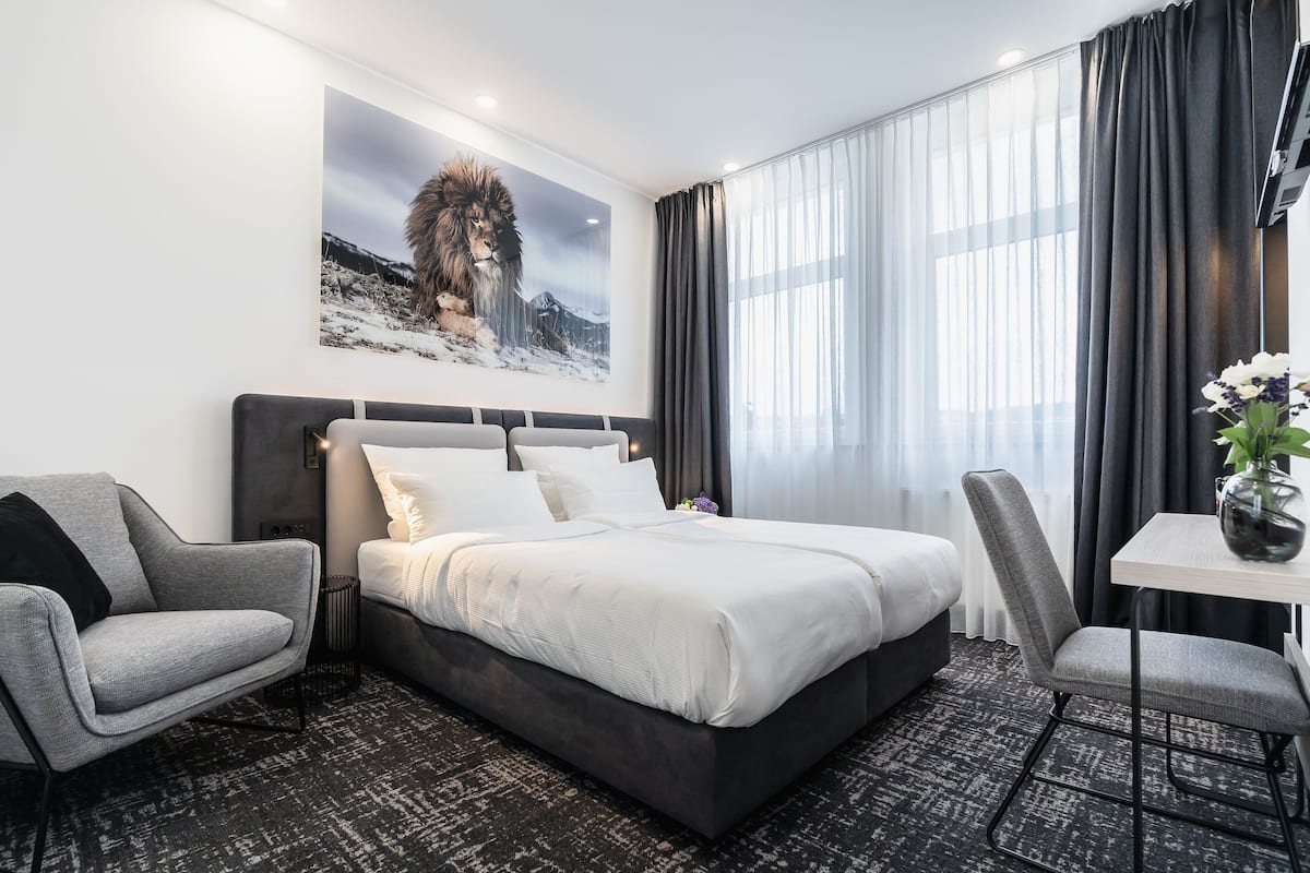 Moderni hotellihuone, jossa on suuri sänky, harmaa nojatuoli, pieni pöytä maljakolla ja seinällä leijonan kuva.