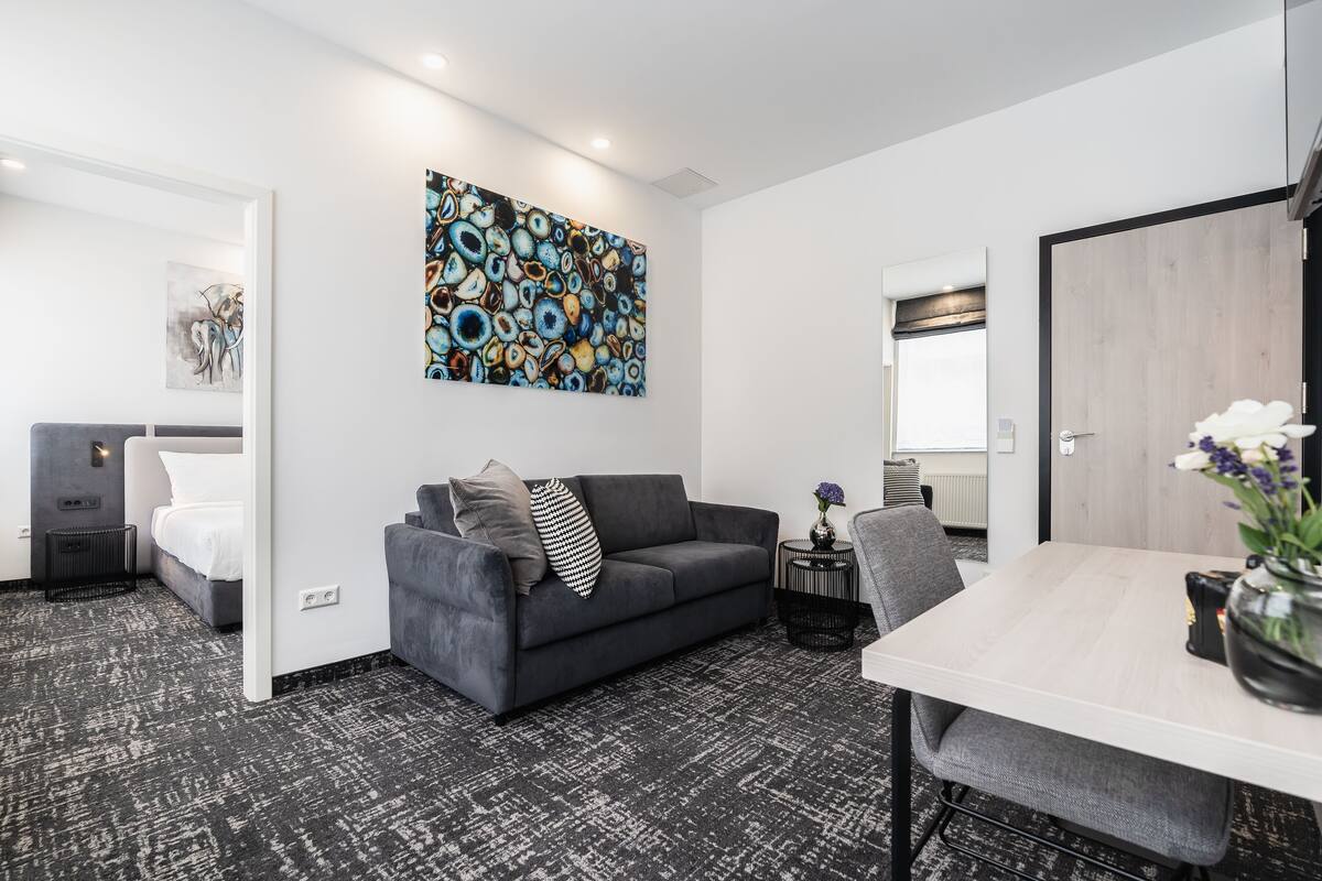 Moderni hotellihuone, jossa on harmaa sohva, pieni pöytä ja suuri abstrakti maalaus seinällä.
