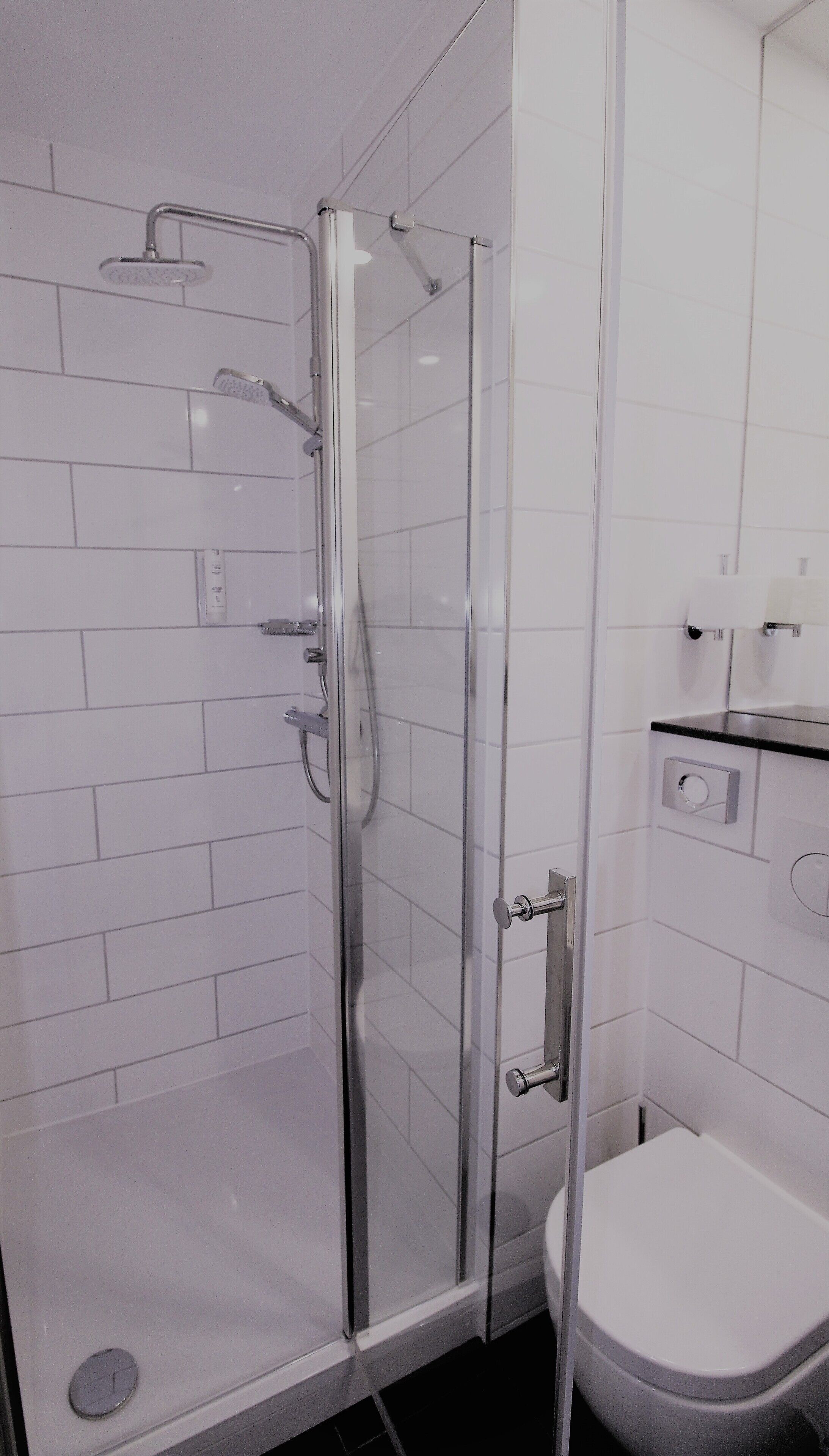Een moderne badkamer met een douchecabine van glas, witte betegelde wanden, een toilet en een klein aanrecht met een spiegel.