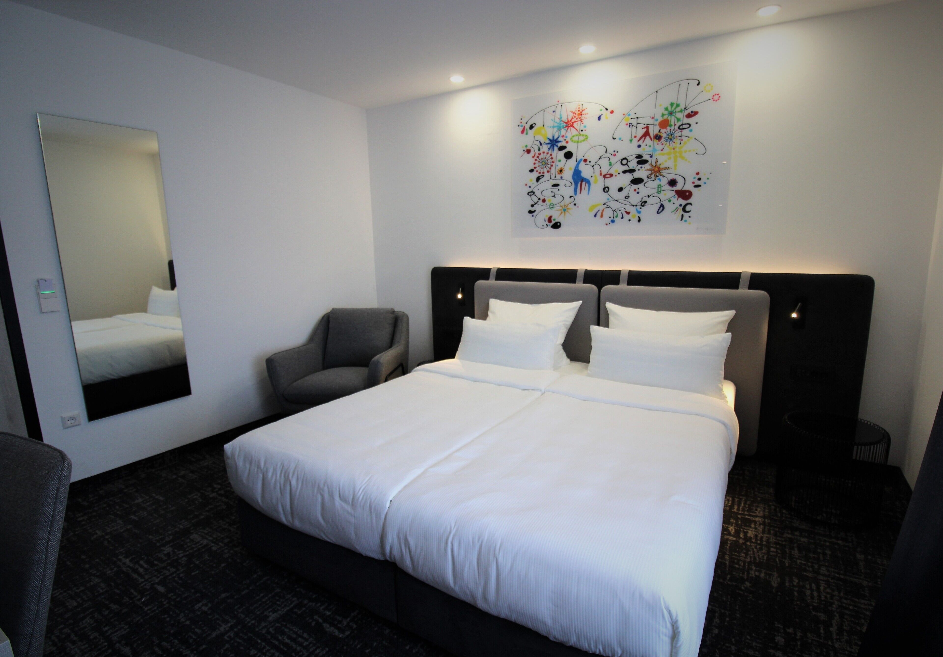 A hotel room with a large bed, a chair, a mirror, and a wall art piece.