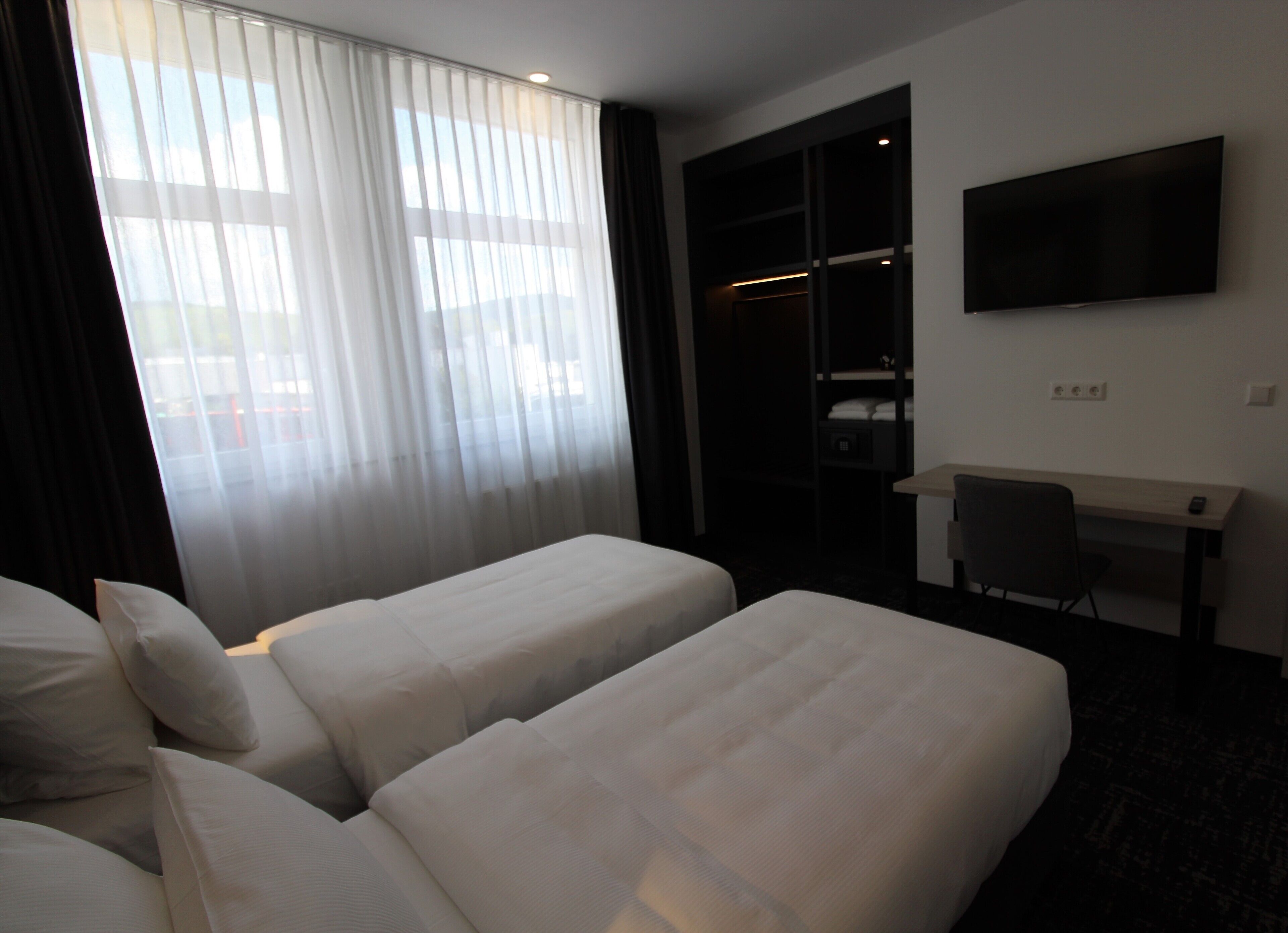 Quarto de hotel com duas camas de solteiro, uma escrivaninha, uma cadeira, uma televisão e um guarda-roupa.