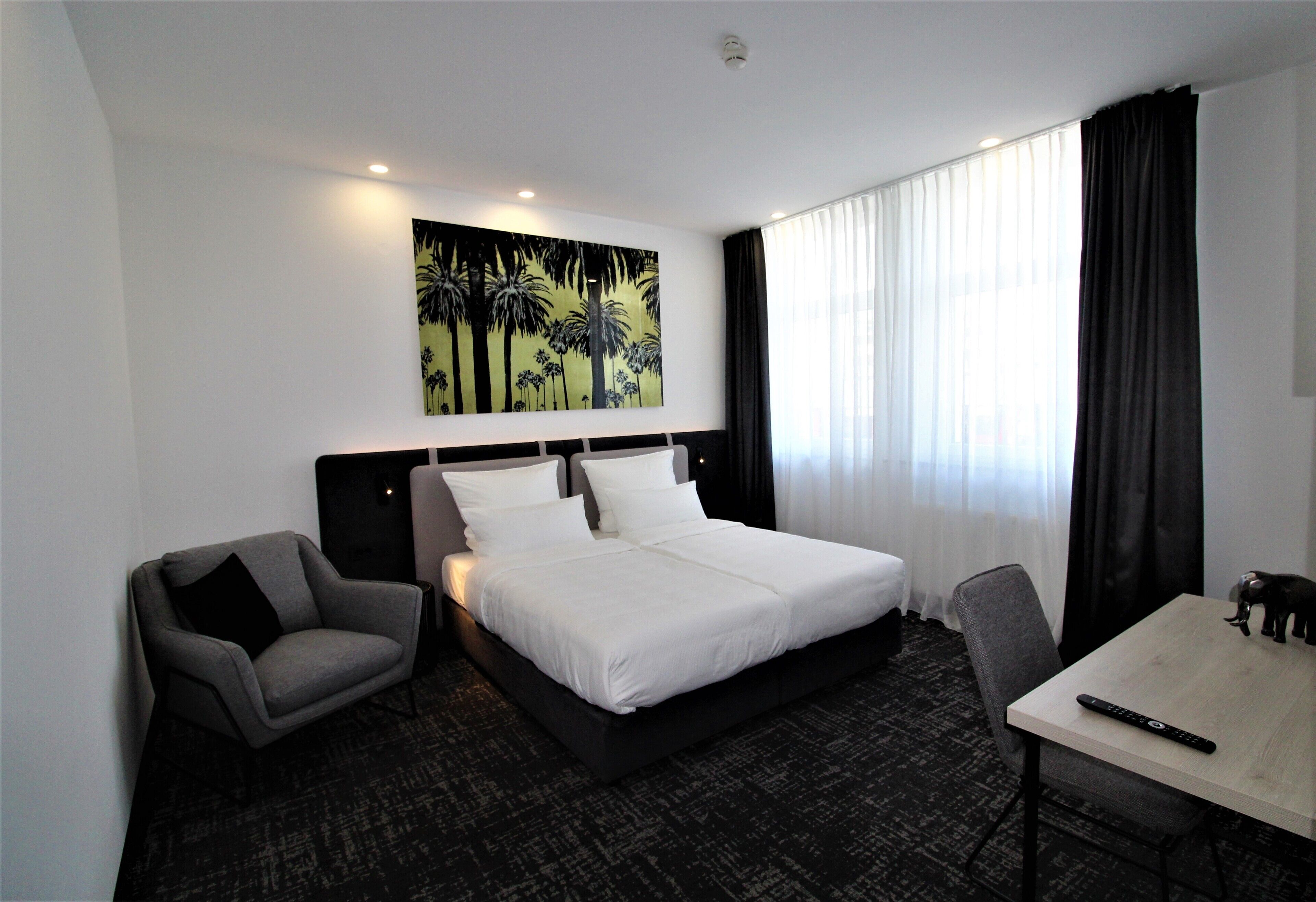 A hotel room with a bed, a chair, a desk, and a painting on the wall.