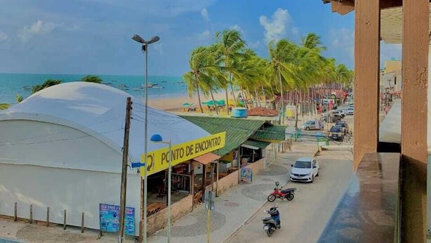 A coastal street with a white car parked on the side, a yellow sign reading "PONTO DE ENCONTRO", and a white tent-like structure.