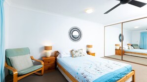 Een slaapkamer met een bed, een stoel, een nachtkastje met een lamp, een spiegel en een plafondventilator.
