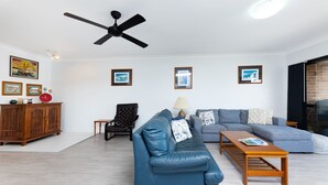 Een woonkamer met een blauwe bank, een houten salontafel, een zwarte leren fauteuil en een plafondventilator.