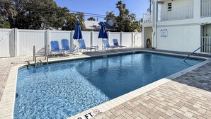 A rectangular pool with a 5 ft depth marker, surrounded by a white fence, blue lounge chairs, and umbrellas.
