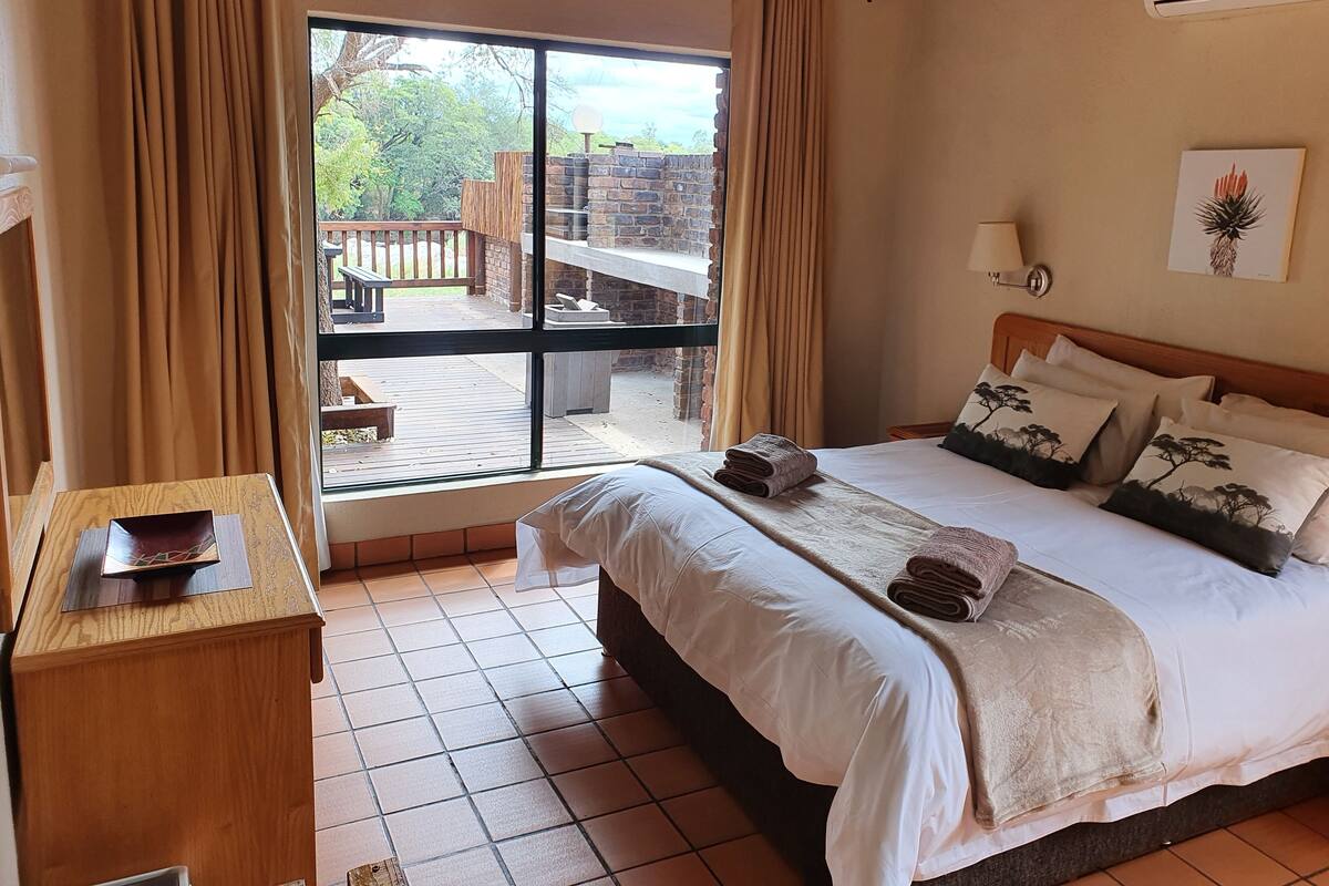 Une chambre avec un grand lit, un bureau en bois, une fenêtre avec des rideaux et une vue sur une terrasse extérieure.