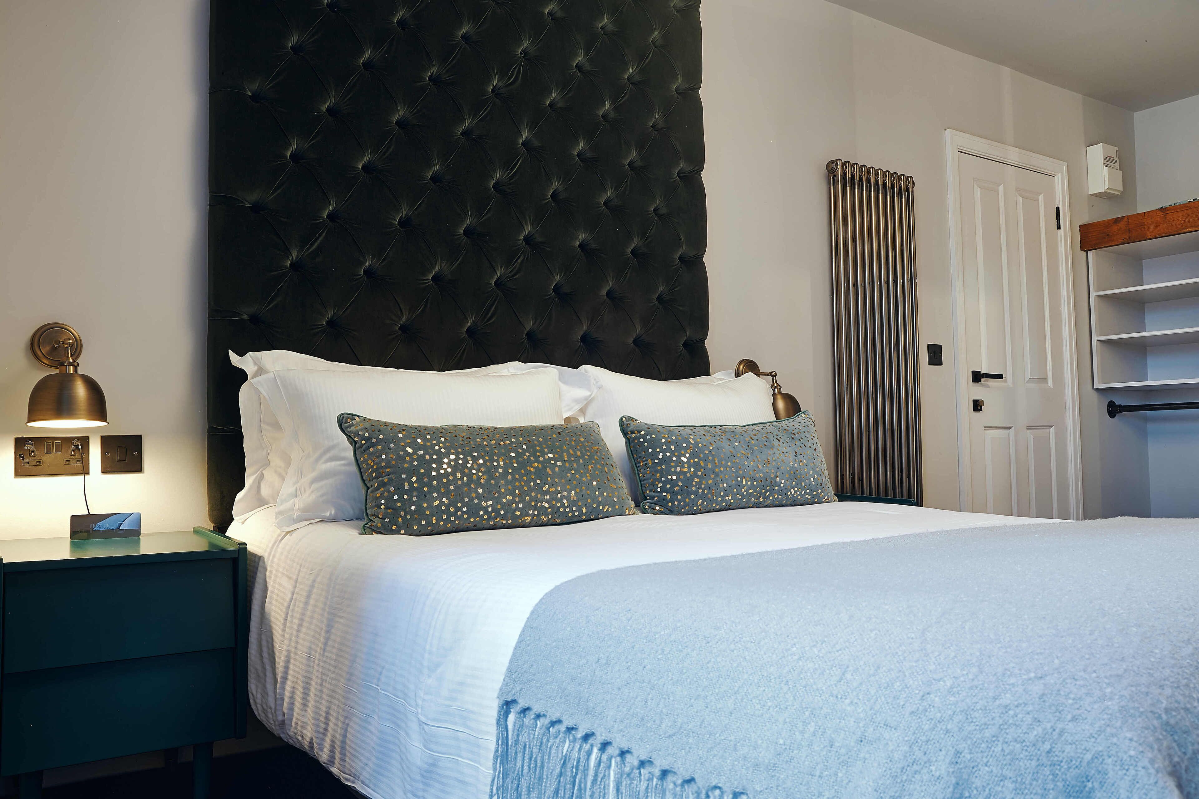 A neatly made bed with a tufted headboard, a bedside table with a lamp, and a white door with a radiator.