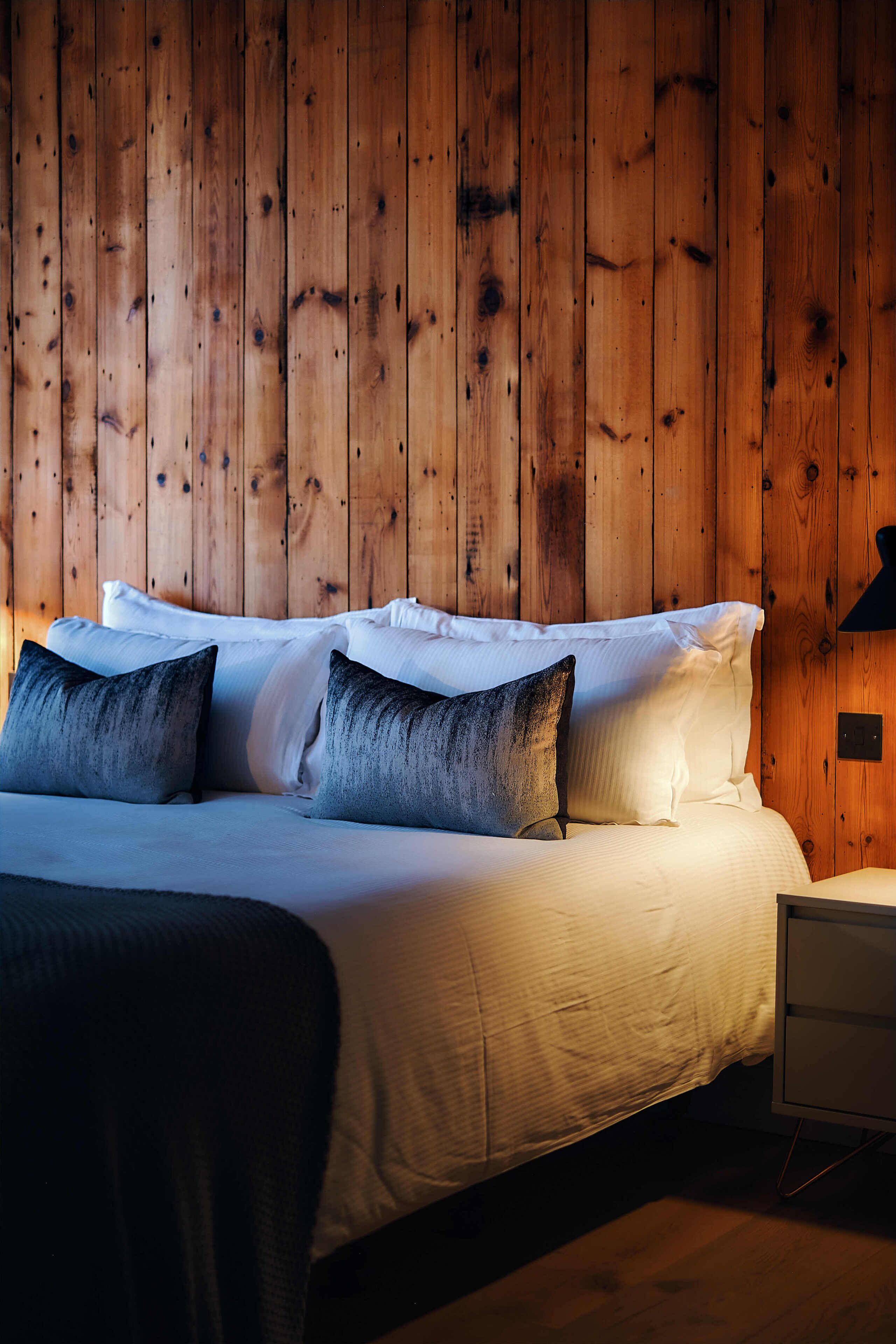 A bed with white bedding and dark pillows, a bedside table, and a wooden wall.