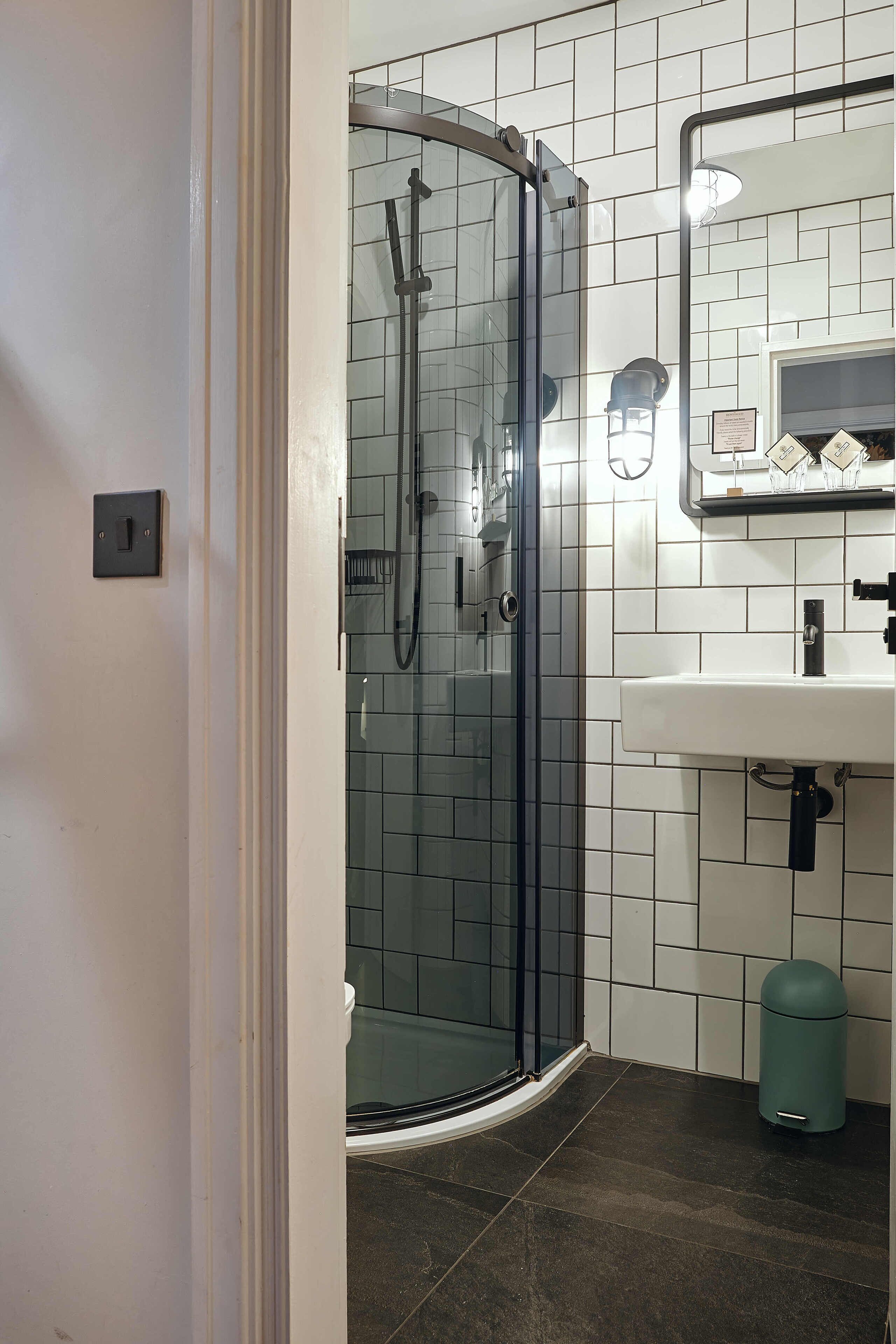 Un baño moderno con ducha de vidrio, paredes de azulejos blancos, lavamanos y un cubo de basura.