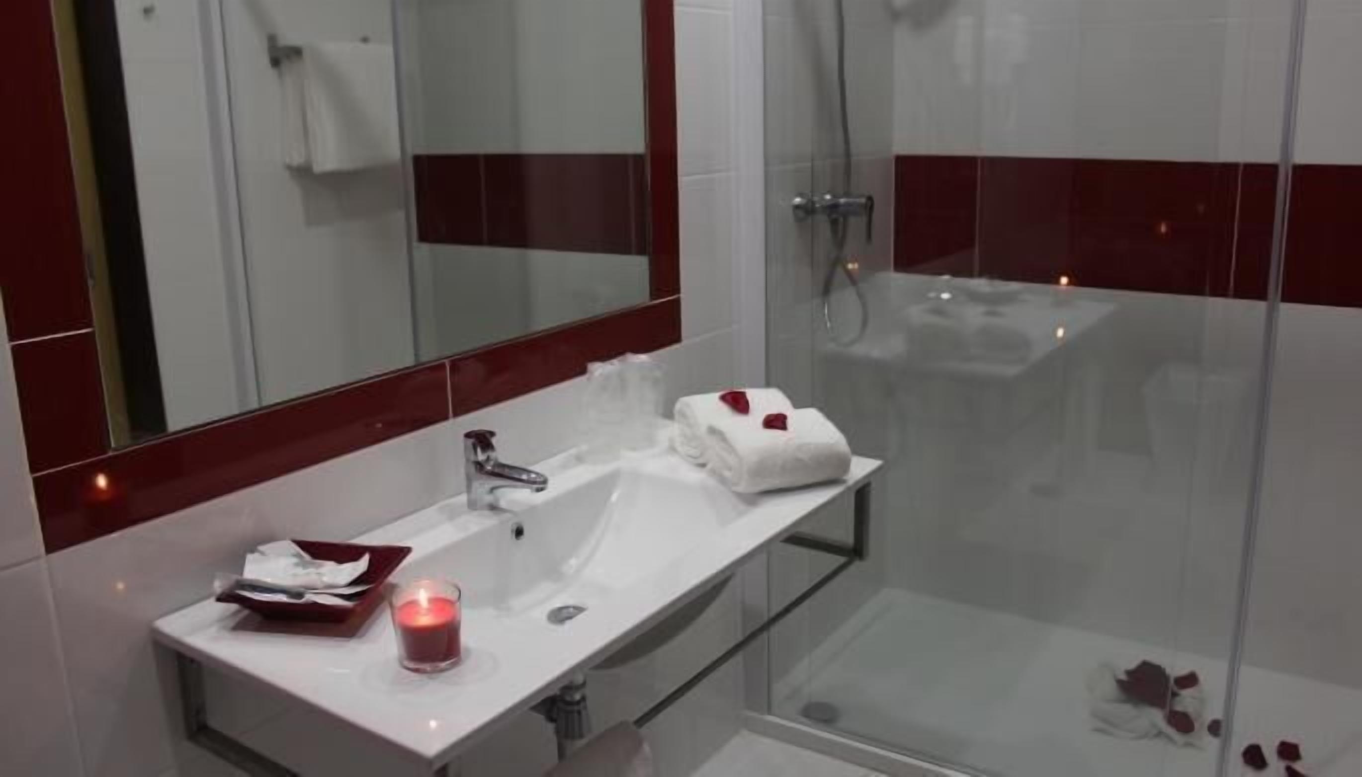 A modern bathroom with a glass shower enclosure, a white sink with a mirror above it, and a small table with a candle and a towel.